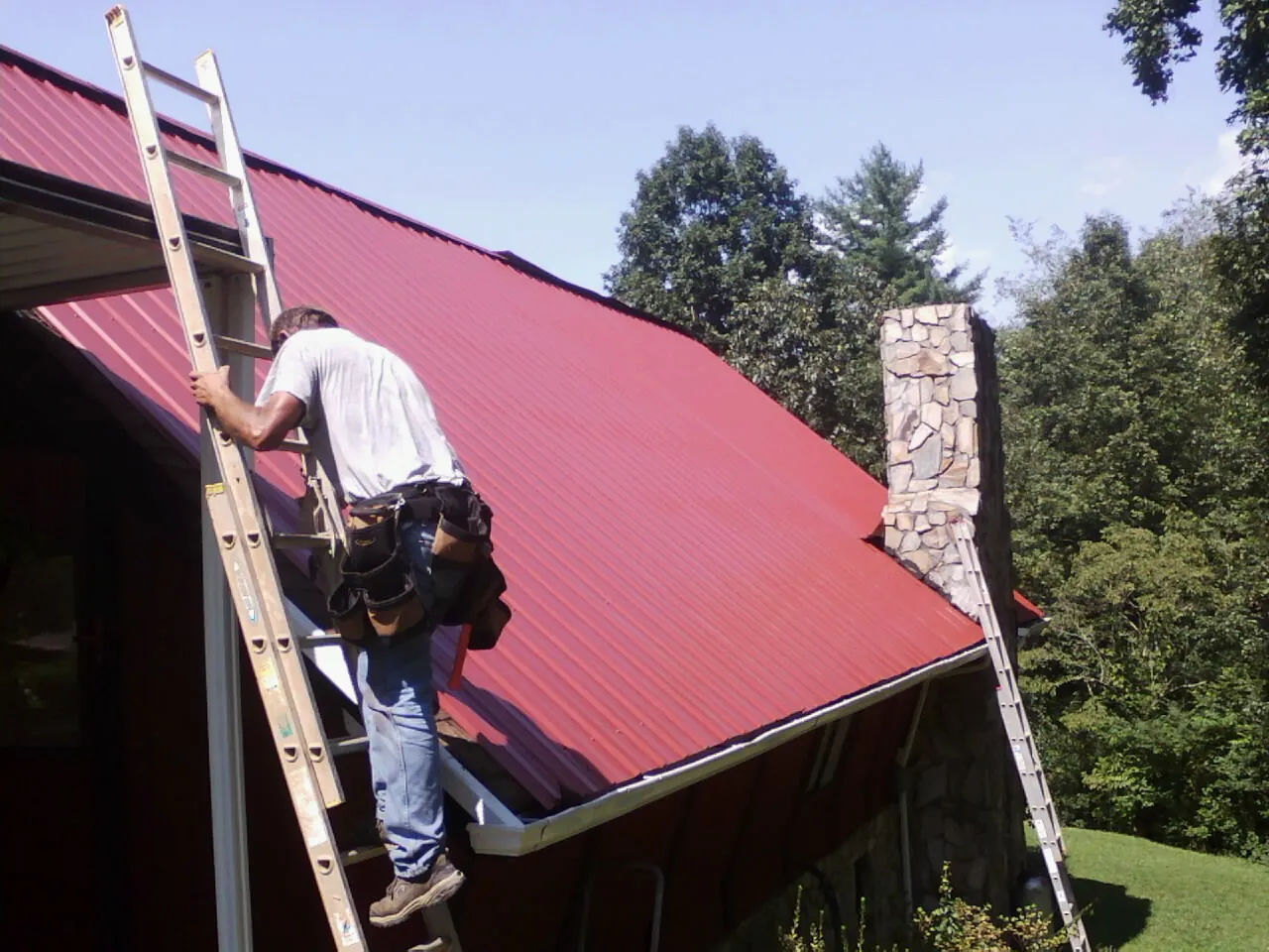 Roofer on a ladder beside a red metal roof and stone chimney — Soffit & Fascia Repair by Roofing Brothers in Mount Freedom